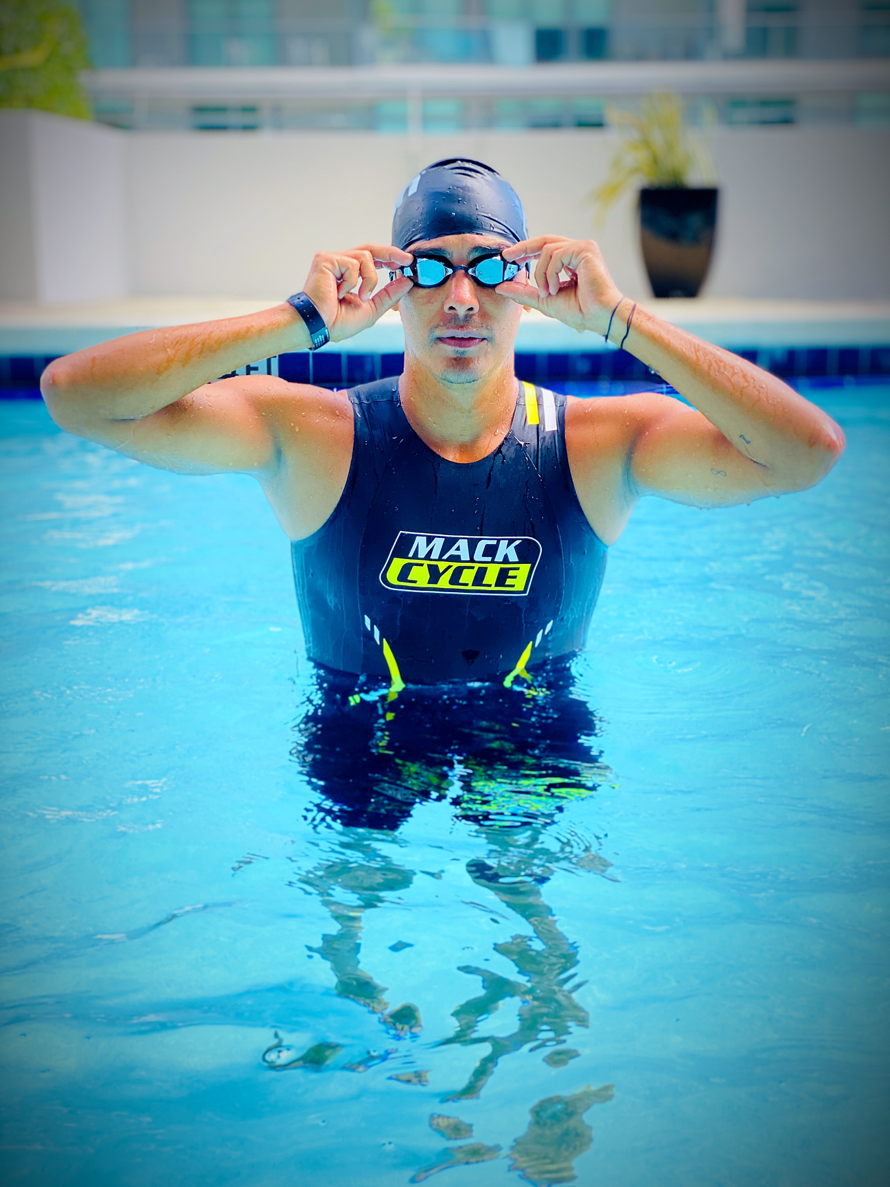 Man (Manuel Novoa) in swimming pool holds on to goggles on his face while emerging from the water wearing the mack cycle triathlon swim skin