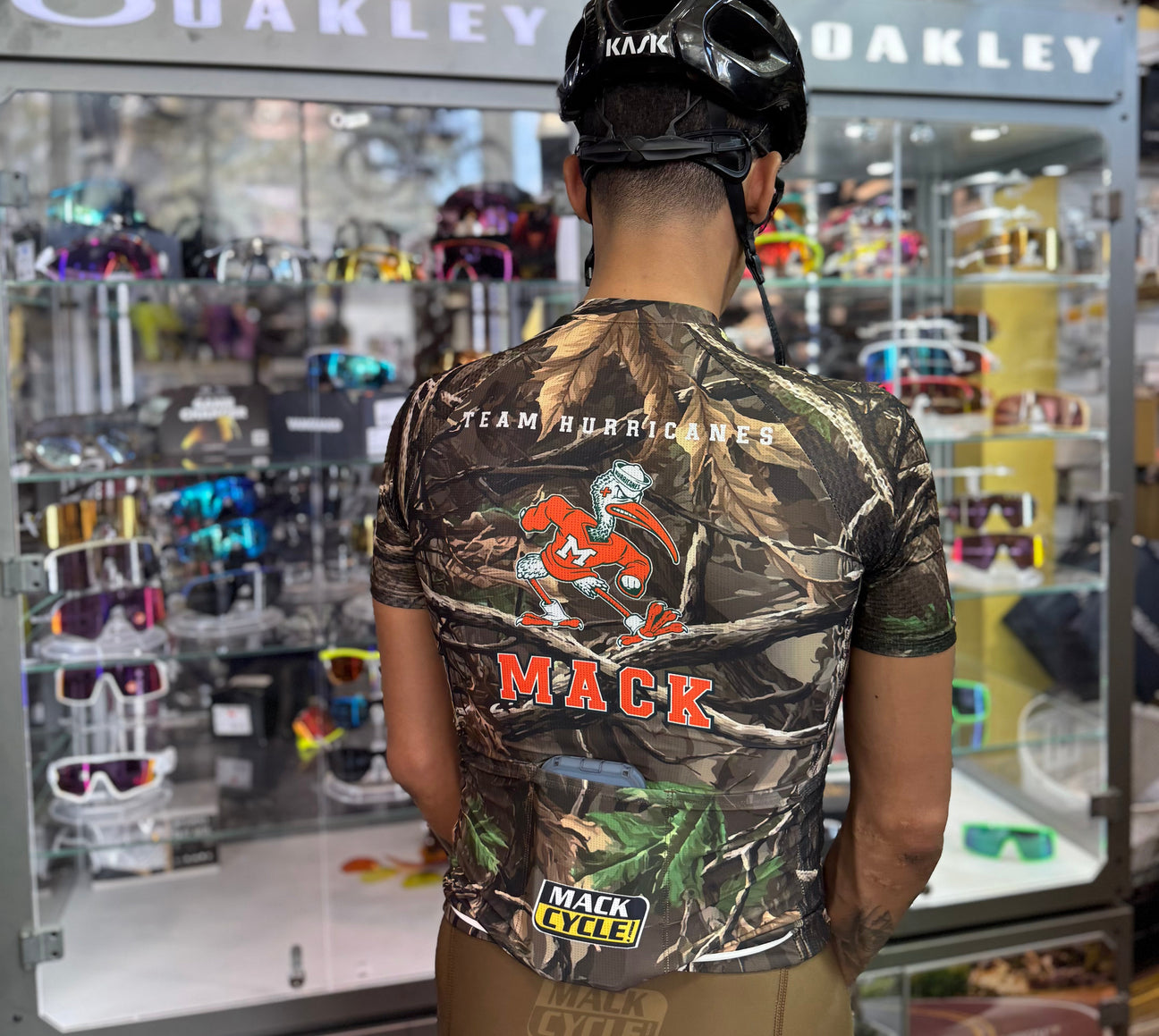 Michael Lum Standing in front of the Oakley Case inside Mack Cycle while wearing his realtree camo team hurricanes x mack cycle hunting season cycling kit and black kask helmet.