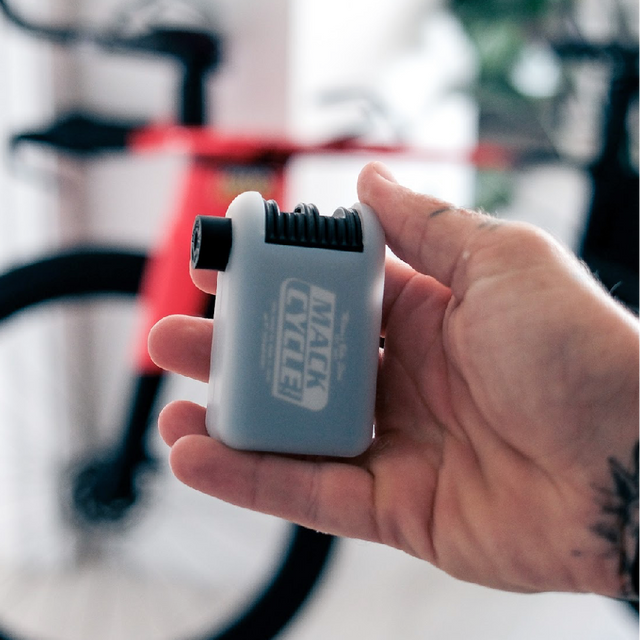 Hand holding a mini electric bicycle pump with a mack cycle logo, blurred background of a room with a BMC speedmachine in red/black